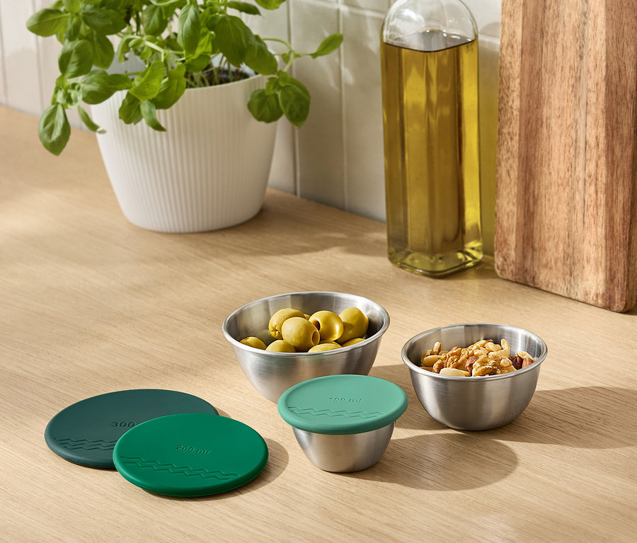 Na blacie kuchennym stoi roślina doniczkowa, butelka oleju, deska do krojenia i metalowe miski z oliwkami i orzechami. Jedna z misek jest przykryta zieloną pokrywką.