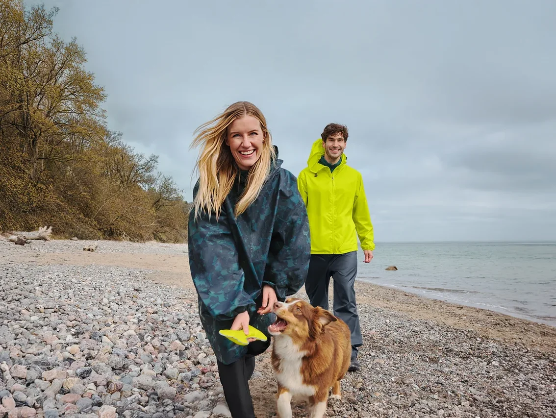 Uśmiechnięta kobieta w ciemnym ponczo przeciwdeszczowym trzyma żółte frisbee, spacerując z brązowo-białym psem po kamienistej plaży. Uśmiechnięty mężczyzna w żółtej kurtce przeciwdeszczowej idzie za nimi, z morzem i zachmurzonym niebem w tle.