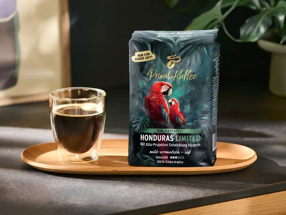 Ciemnozielona torebka kawy „Privat Kaffee Honduras Limited” z dwoma czerwonymi arami i szklana filiżanka czarnej kawy stoją na drewnianej tacy. Na torebce widnieją napisy takie jak „NUR FÜR KURZE ZEIT!” i „100% Tchibo Arabica”. W tle znajduje się zielona roślina.